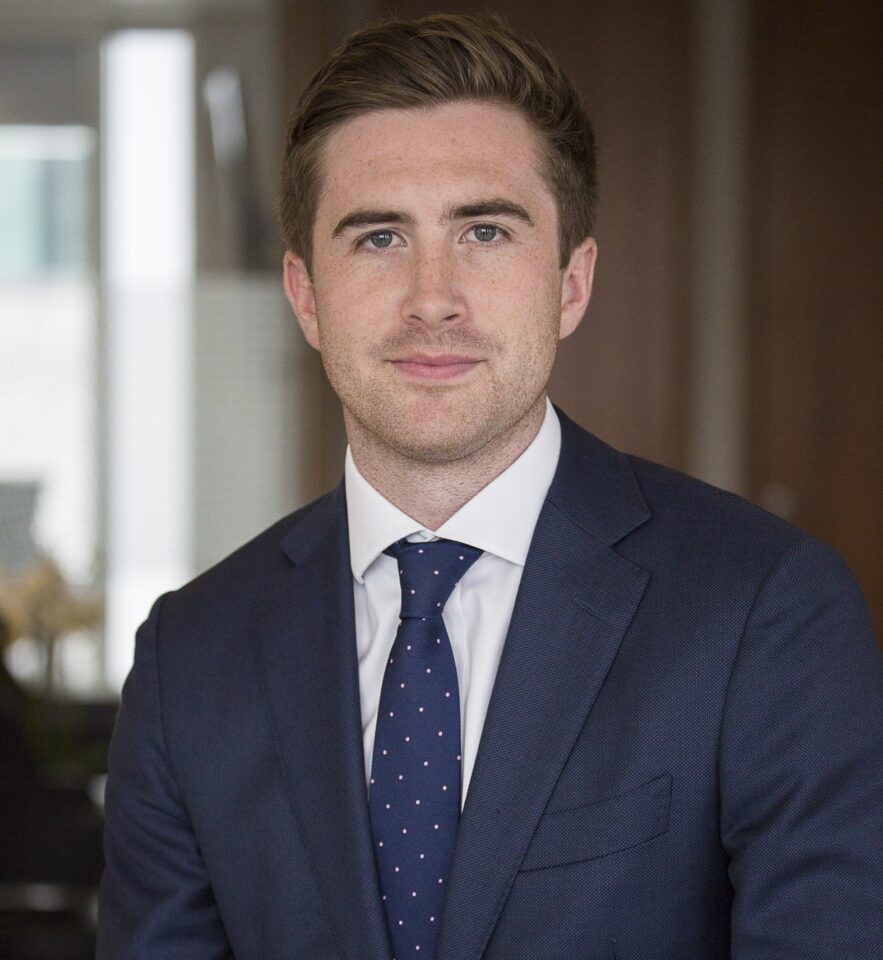 Headshot of a man with light brown hair and blue eyes, wearing a navy suit and a dark blue tie with pink polka dots.