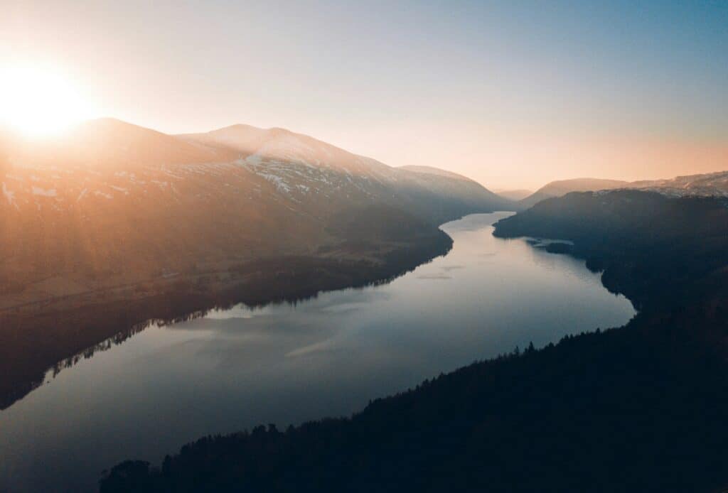 Aerial view of a long, winding lake surrounded by mountains with patches of snow. A bright sunburst illuminates the left side, casting a warm glow over the landscape.