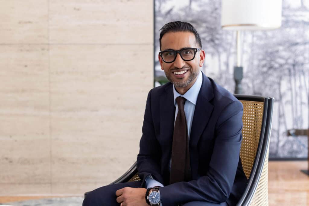 A smiling man in a dark blue suit, light blue shirt, dark brown tie, and black glasses sits in a cane chair. He has a beard and dark hair. The background includes a light-colored tiled wall and a black and white patterned wall with a lamp.