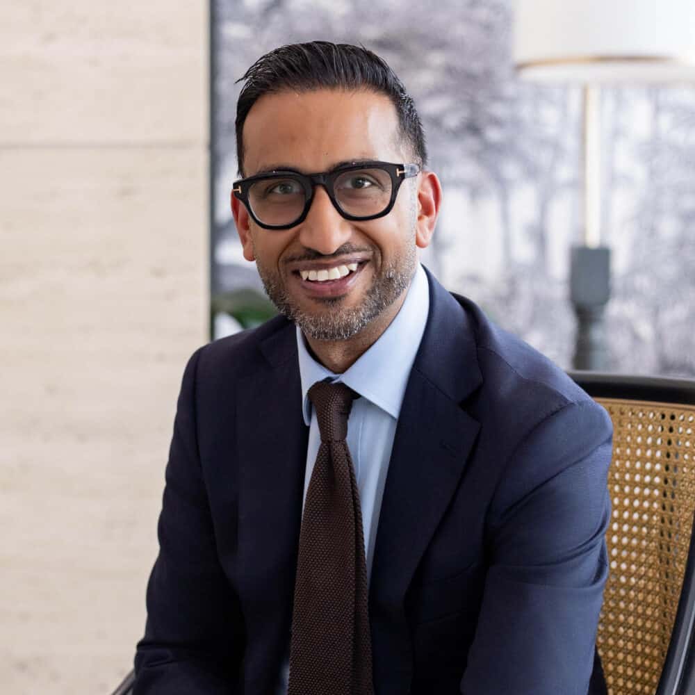 A smiling man in a dark blue suit, light blue shirt, dark brown tie, and black glasses sits in a cane chair. He has a beard and dark hair. The background includes a light-colored tiled wall and a black and white patterned wall with a lamp.
