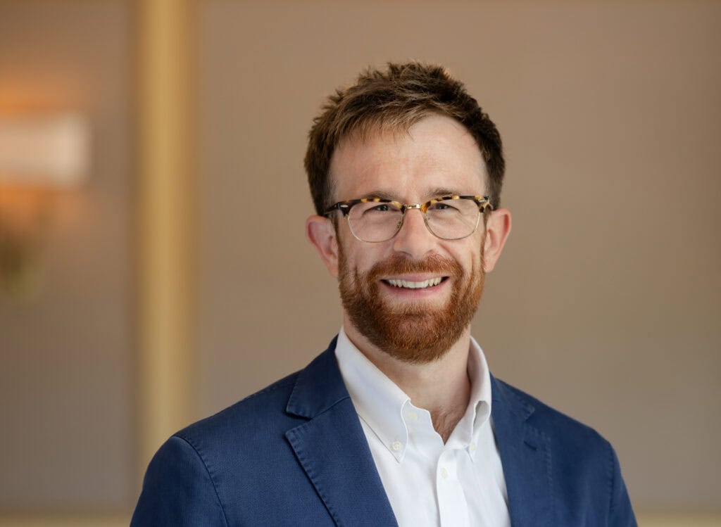 Headshot of a smiling man with a reddish-brown beard and glasses, wearing a blue blazer and white shirt.
