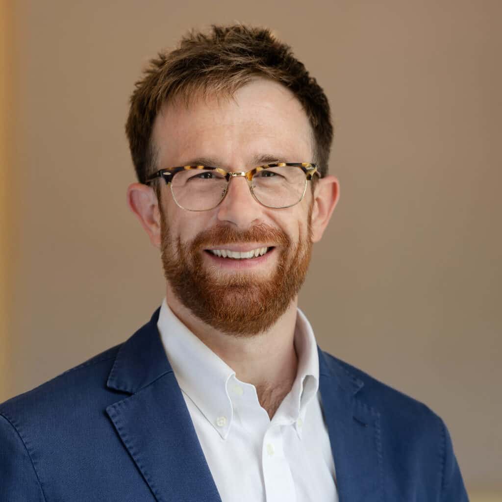 Headshot of a smiling man with a reddish-brown beard and glasses, wearing a blue blazer and white shirt.