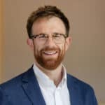 Headshot of a smiling man with a reddish-brown beard and glasses, wearing a blue blazer and white shirt.