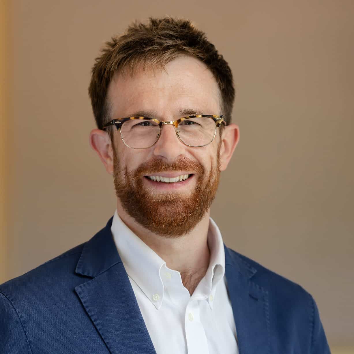Headshot of a smiling man with a reddish-brown beard and glasses, wearing a blue blazer and white shirt.