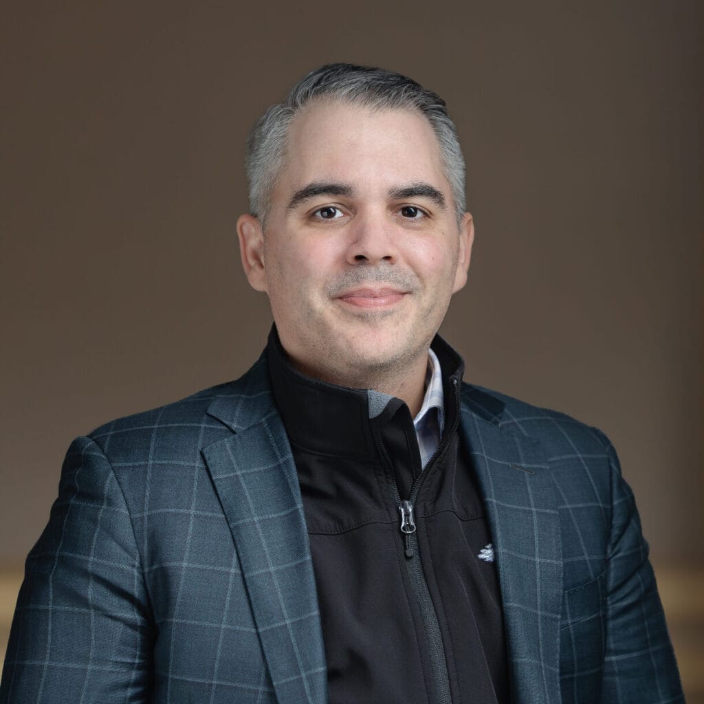 Headshot of a man with graying hair, wearing a plaid suit jacket over a black zip-up, smiling at the camera.