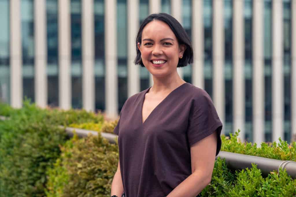 A smiling woman with short dark hair and a brown top stands in front of a modern building and green bushes.
