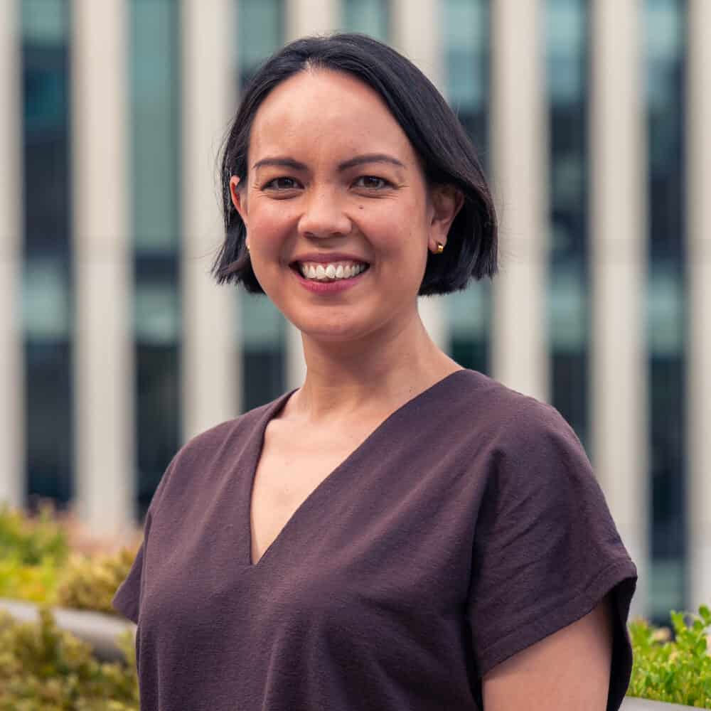 A smiling woman with short dark hair and a brown top stands in front of a modern building and green bushes.