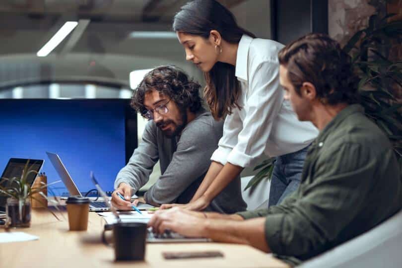 Three colleagues collaborating at a desk in an office. One man writes, a woman observes, and another man types on a laptop.