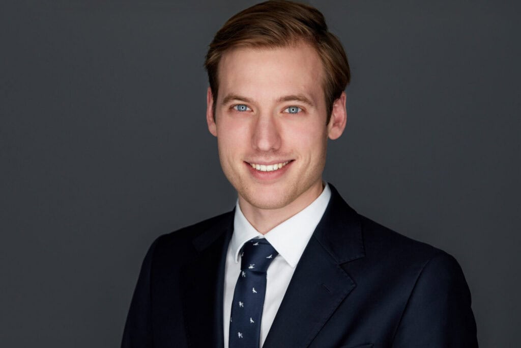 Headshot of a smiling young man with light brown hair and blue eyes, wearing a navy suit and tie against a gray background.