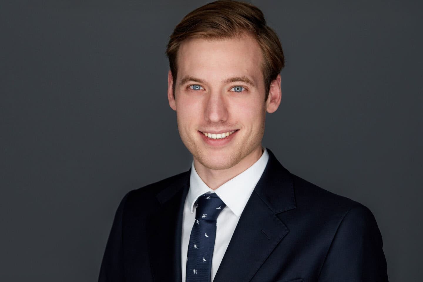 Headshot of a smiling young man with light brown hair and blue eyes, wearing a navy suit and tie against a gray background.