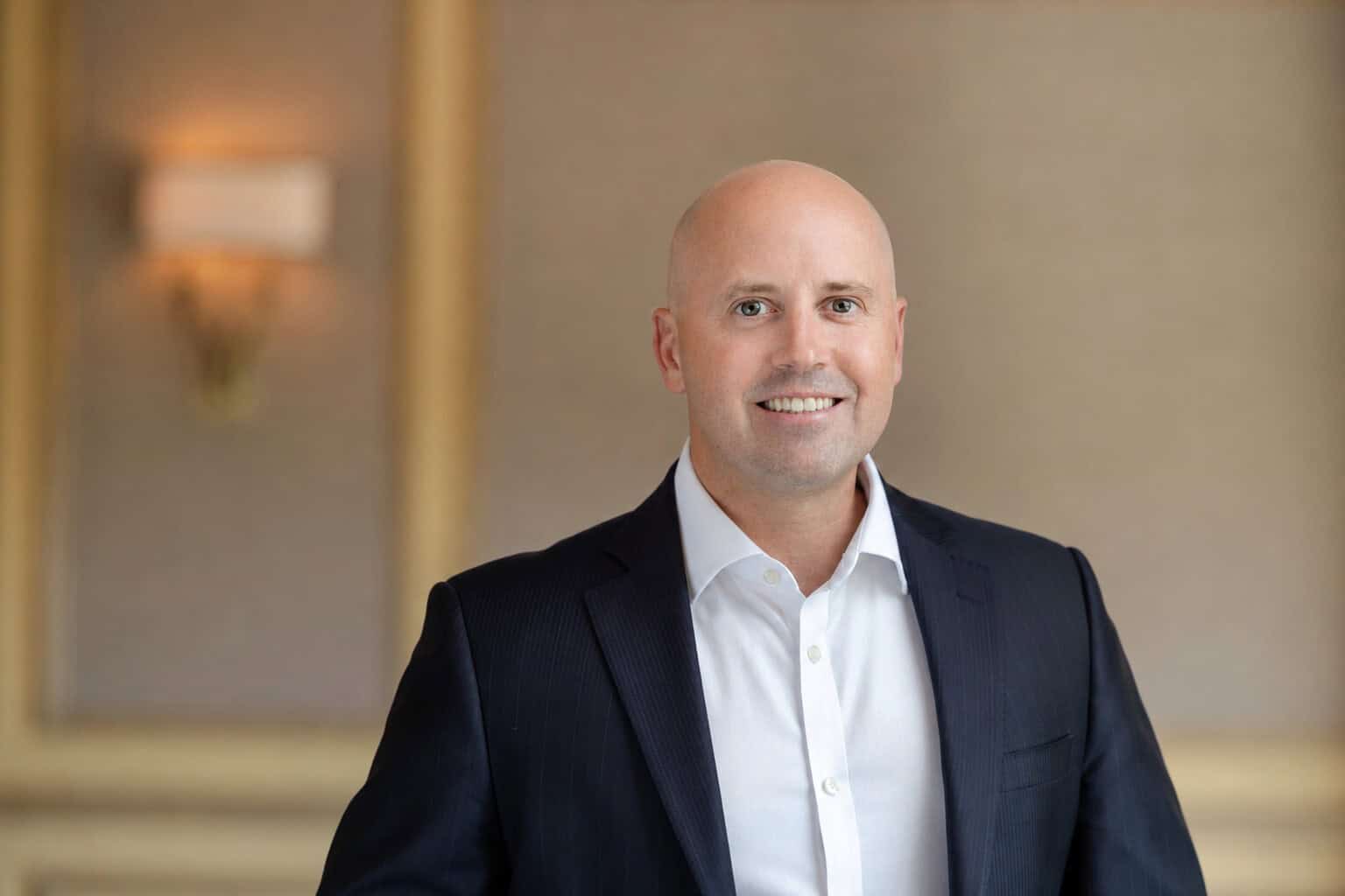 A bald man with light skin and light eyes smiles at the camera, wearing a dark suit jacket and white shirt.