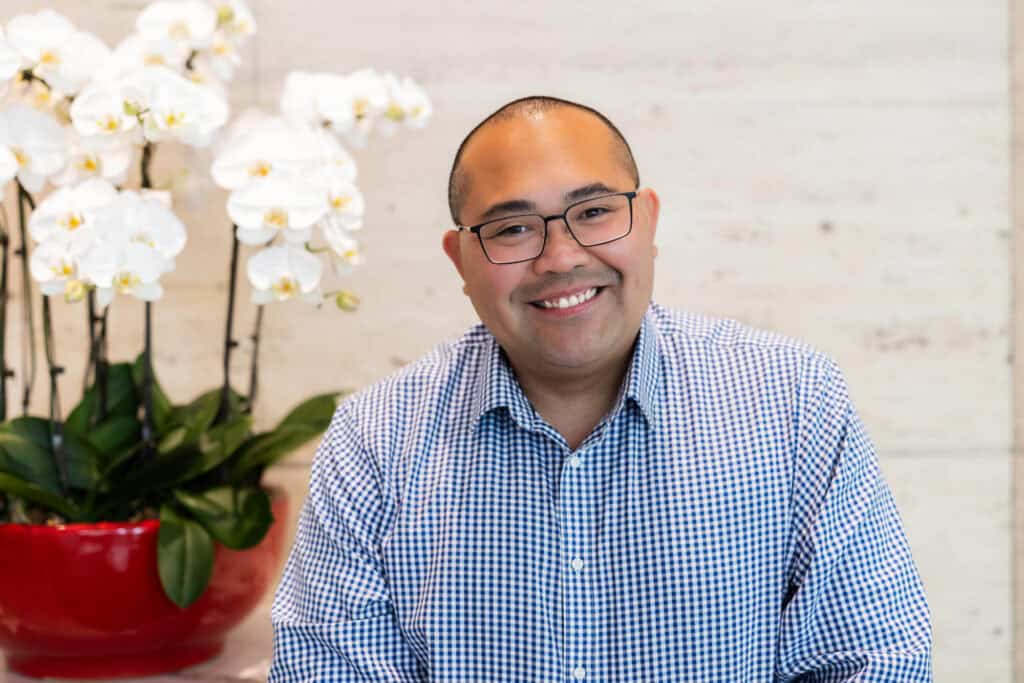 A man with glasses and a blue gingham shirt smiles in front of white orchids.