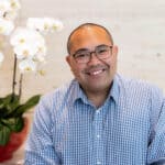 A man with glasses and a blue gingham shirt smiles in front of white orchids.