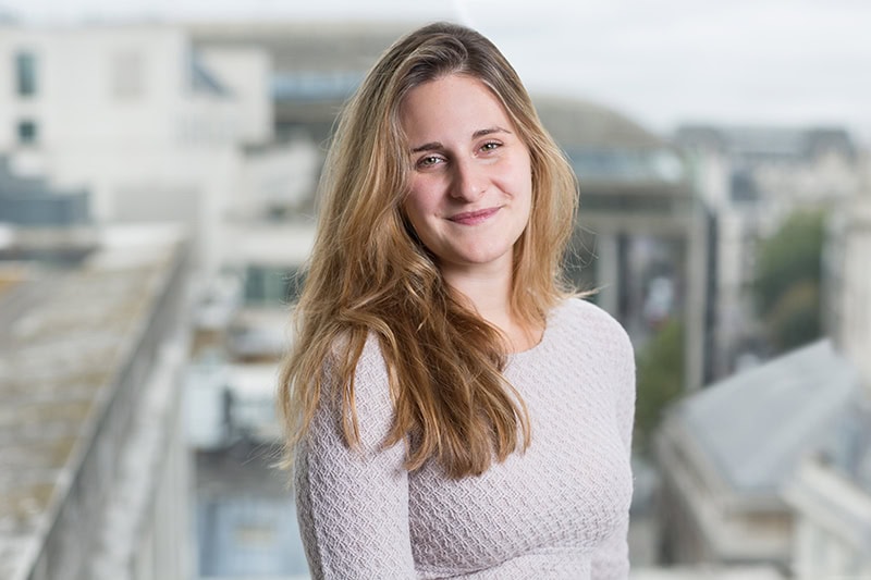 Headshot of a smiling woman with long light brown hair and a light gray top, with a blurred city background.