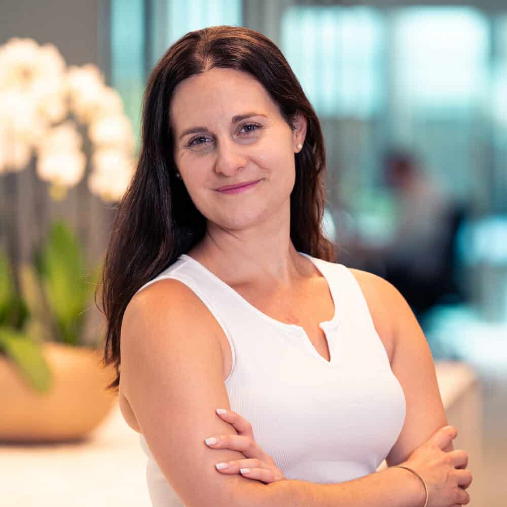 A woman with dark hair and a white top stands with crossed arms, smiling at the camera.