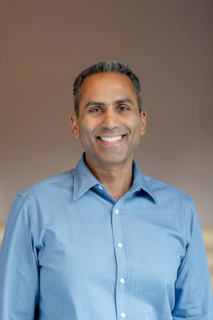 Headshot of a smiling man with dark hair and a light blue shirt against a blurred brown background.
