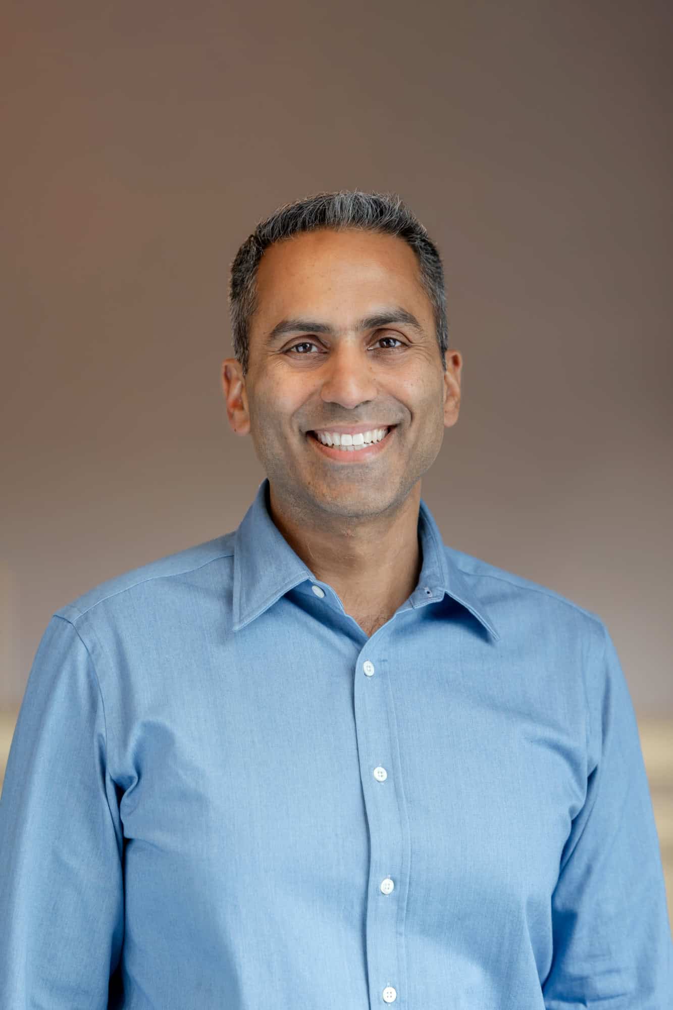 Headshot of a smiling man with dark hair and a light blue shirt against a blurred brown background.