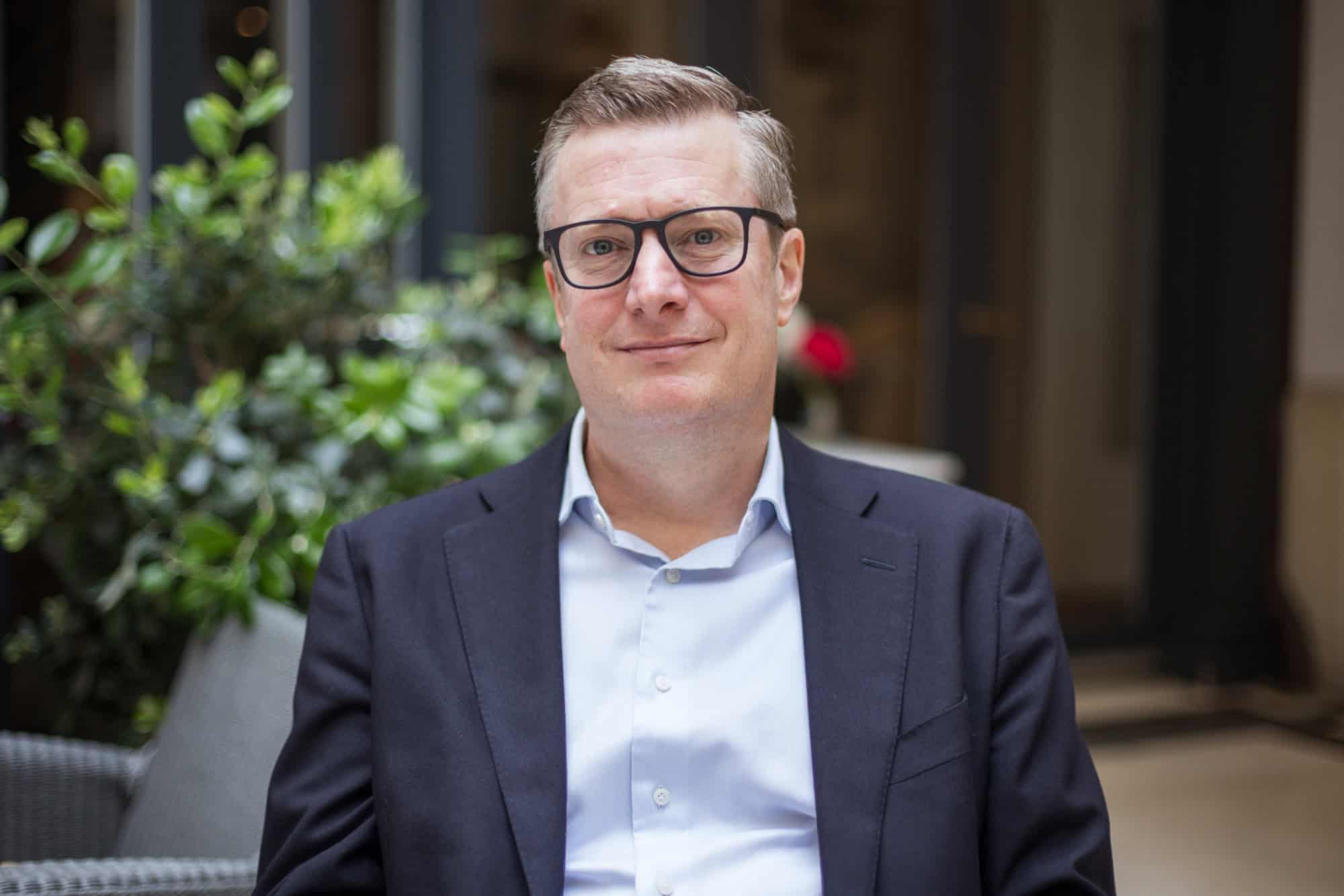 A man with glasses and a suit jacket smiles directly at the camera.