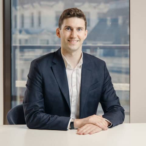A man in a dark suit and white shirt smiles while seated at a white table, with a blurry office window in the background.