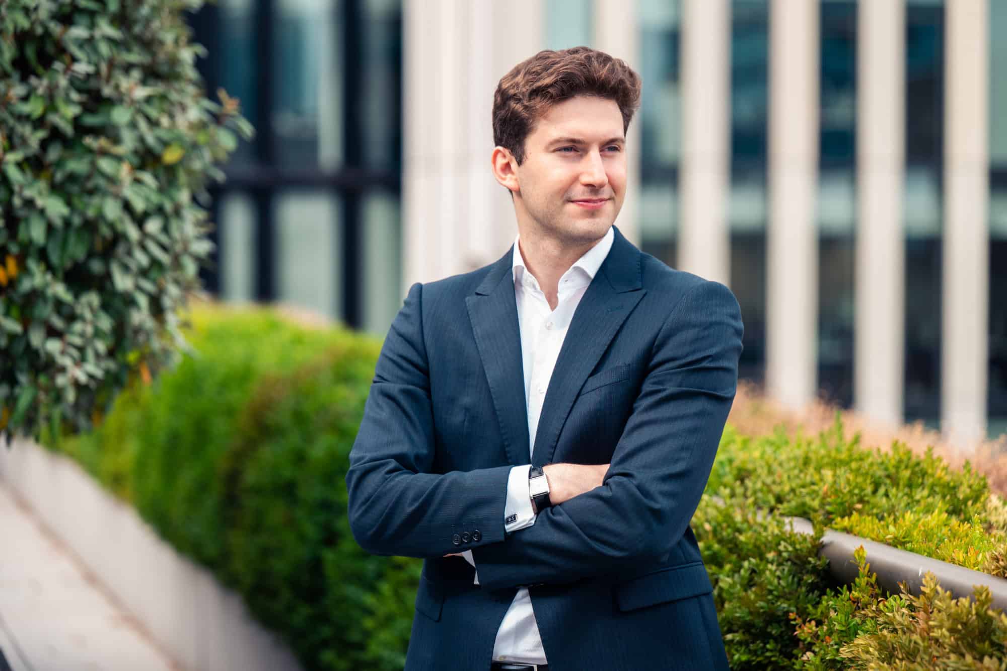 A young man in a dark blue suit and white shirt, with arms crossed, stands outdoors smiling slightly. Green foliage and a blurred modern building are in the background.