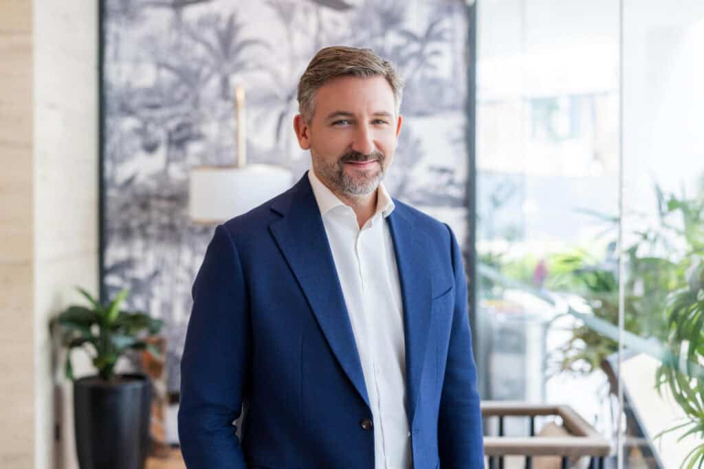 A man with graying hair and a beard, wearing a blue suit jacket and white shirt, smiles at the camera in an indoor setting with a tropical-themed background.