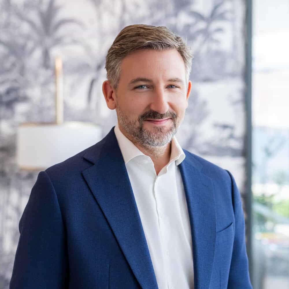 A man with graying hair and a beard, wearing a blue suit jacket and white shirt, smiles at the camera in an indoor setting with a tropical-themed background.