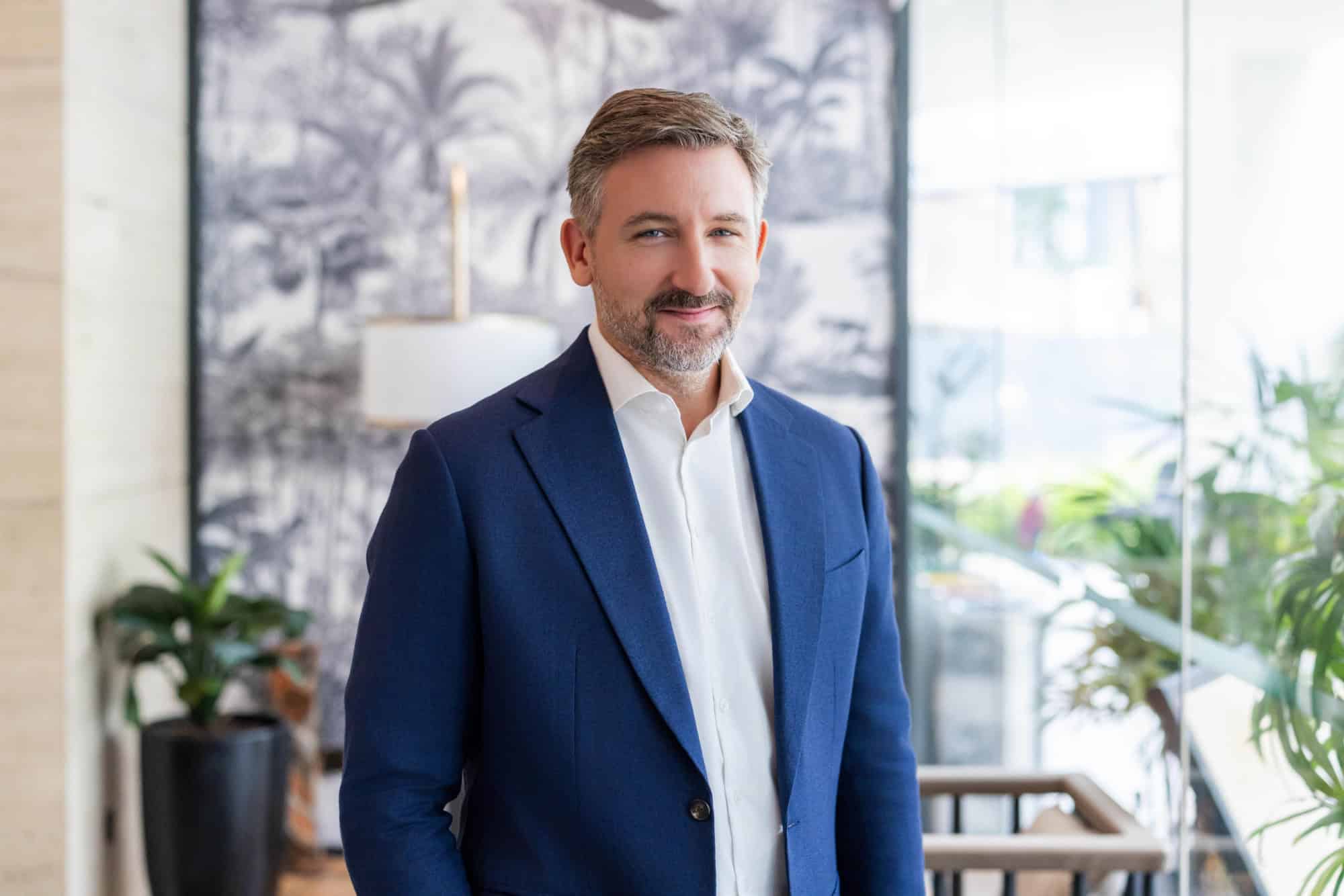 A man with graying hair and a beard, wearing a blue suit jacket and white shirt, smiles at the camera in an indoor setting with a tropical-themed background.