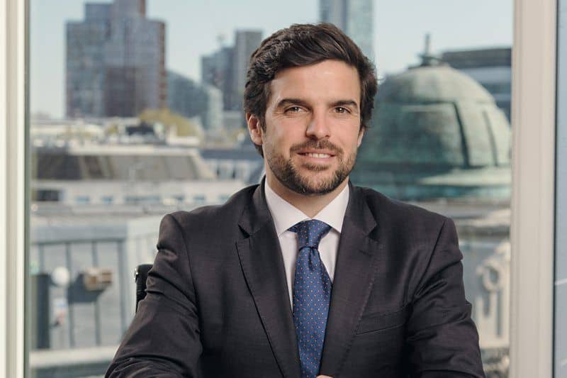 A man with dark hair and a beard, wearing a suit and tie, smiles at the camera in front of a city window.