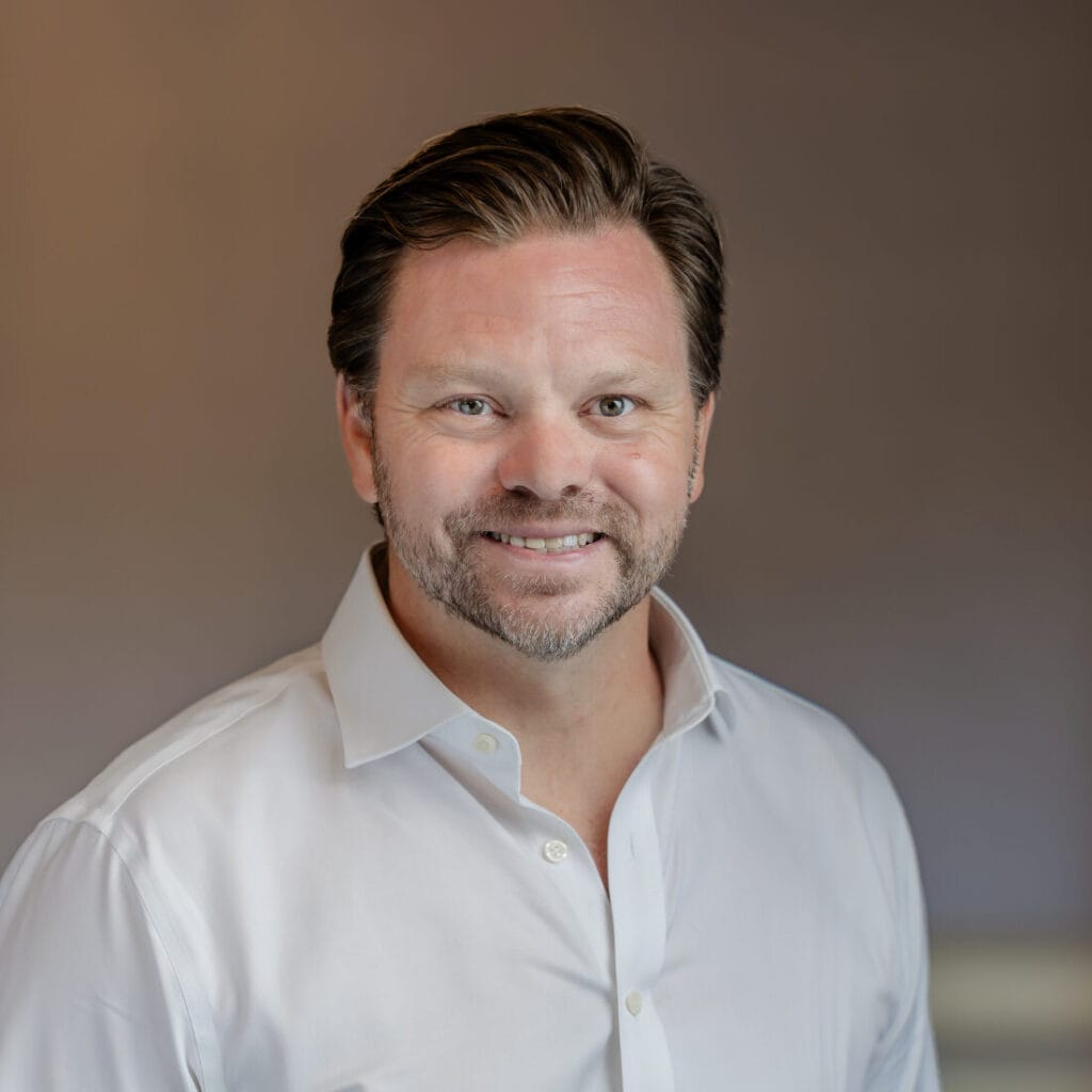 Headshot of a smiling man with dark hair and a beard, wearing a white shirt, against a blurred background with a warm light.