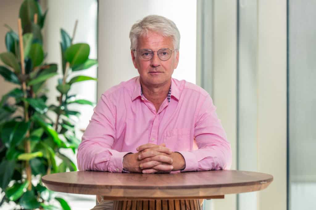 A man with gray hair and glasses in a pink shirt, sitting at a wooden table with hands clasped.