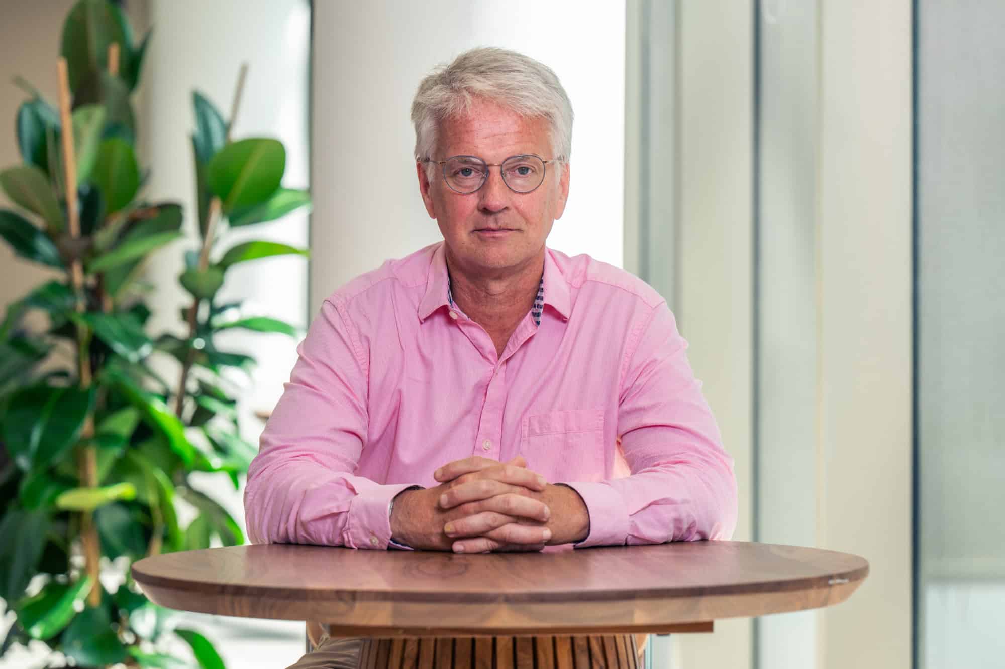 A man with gray hair and glasses in a pink shirt, sitting at a wooden table with hands clasped.