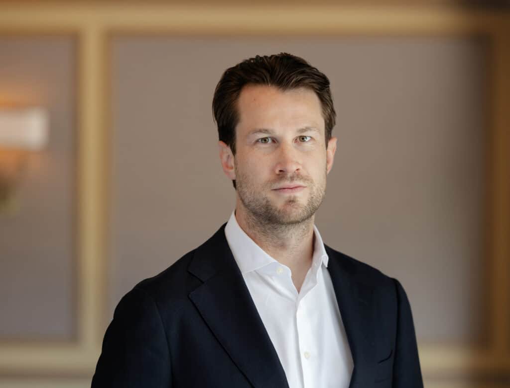 Headshot of a man with brown hair and light eyes, wearing a dark suit jacket and white shirt, looking forward.