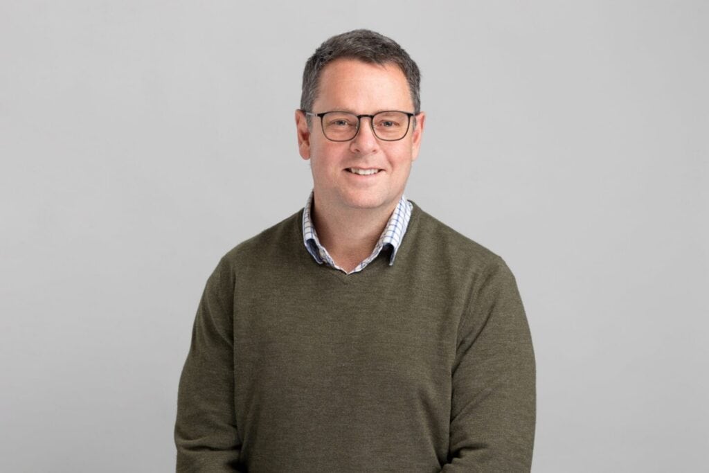 Headshot of a smiling man with glasses, wearing a green sweater over a checkered shirt, against a gray background.