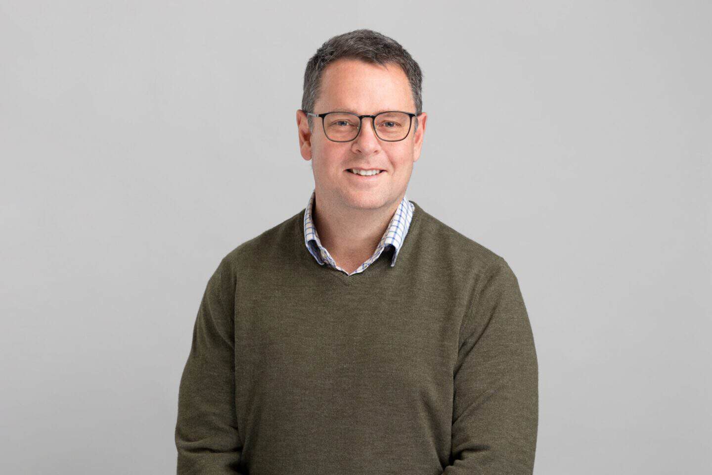 Headshot of a smiling man with glasses, wearing a green sweater over a checkered shirt, against a gray background.