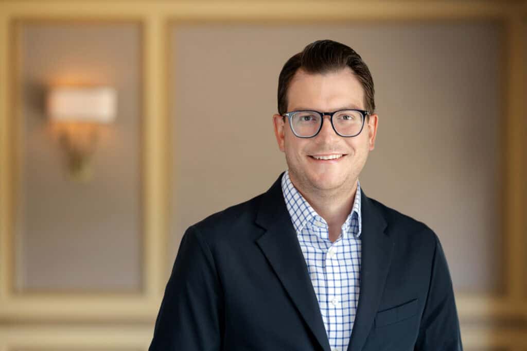 Headshot of a smiling man with dark hair and glasses, wearing a dark blazer and a blue and white checkered shirt.