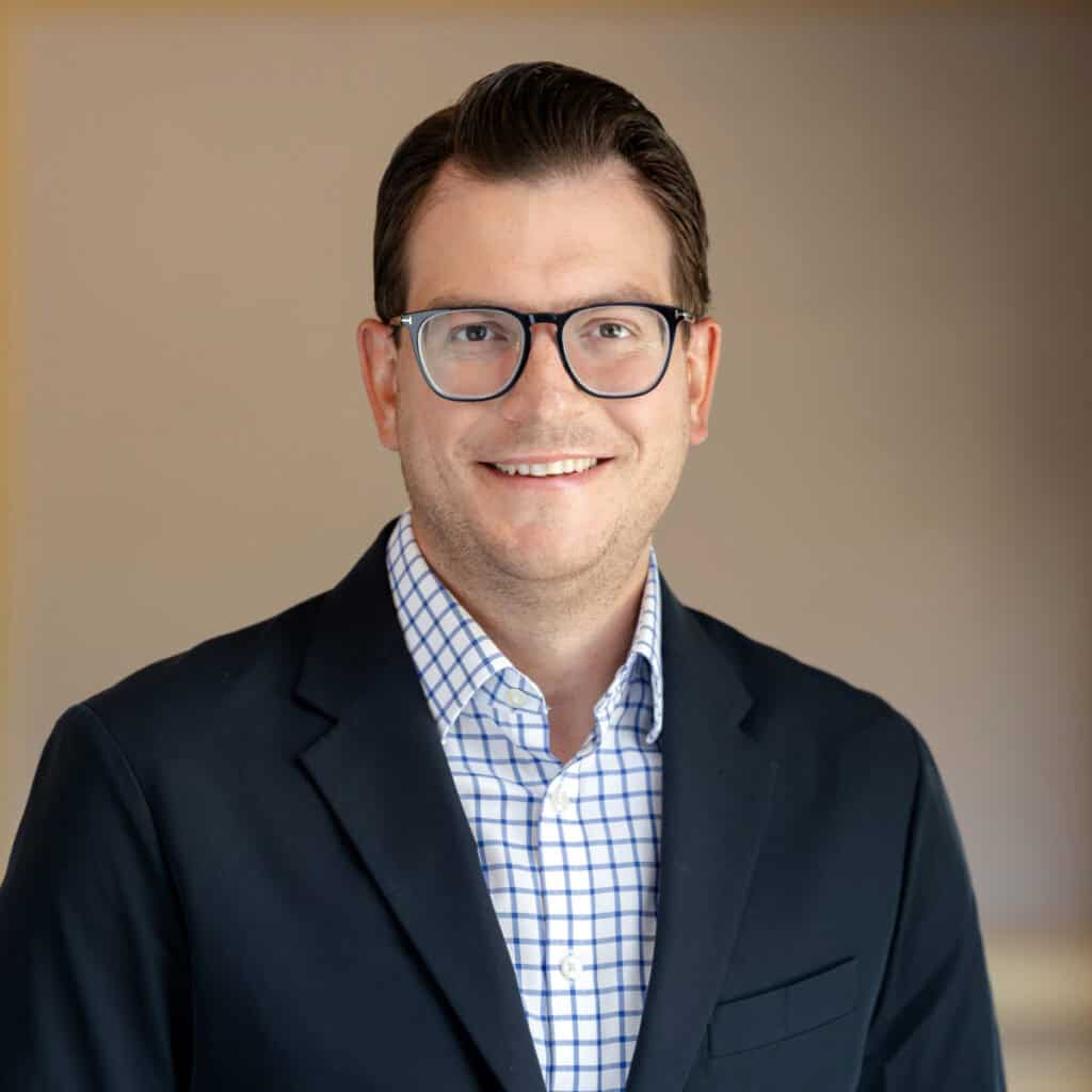 Headshot of a smiling man with dark hair and glasses, wearing a dark blazer and a blue and white checkered shirt.
