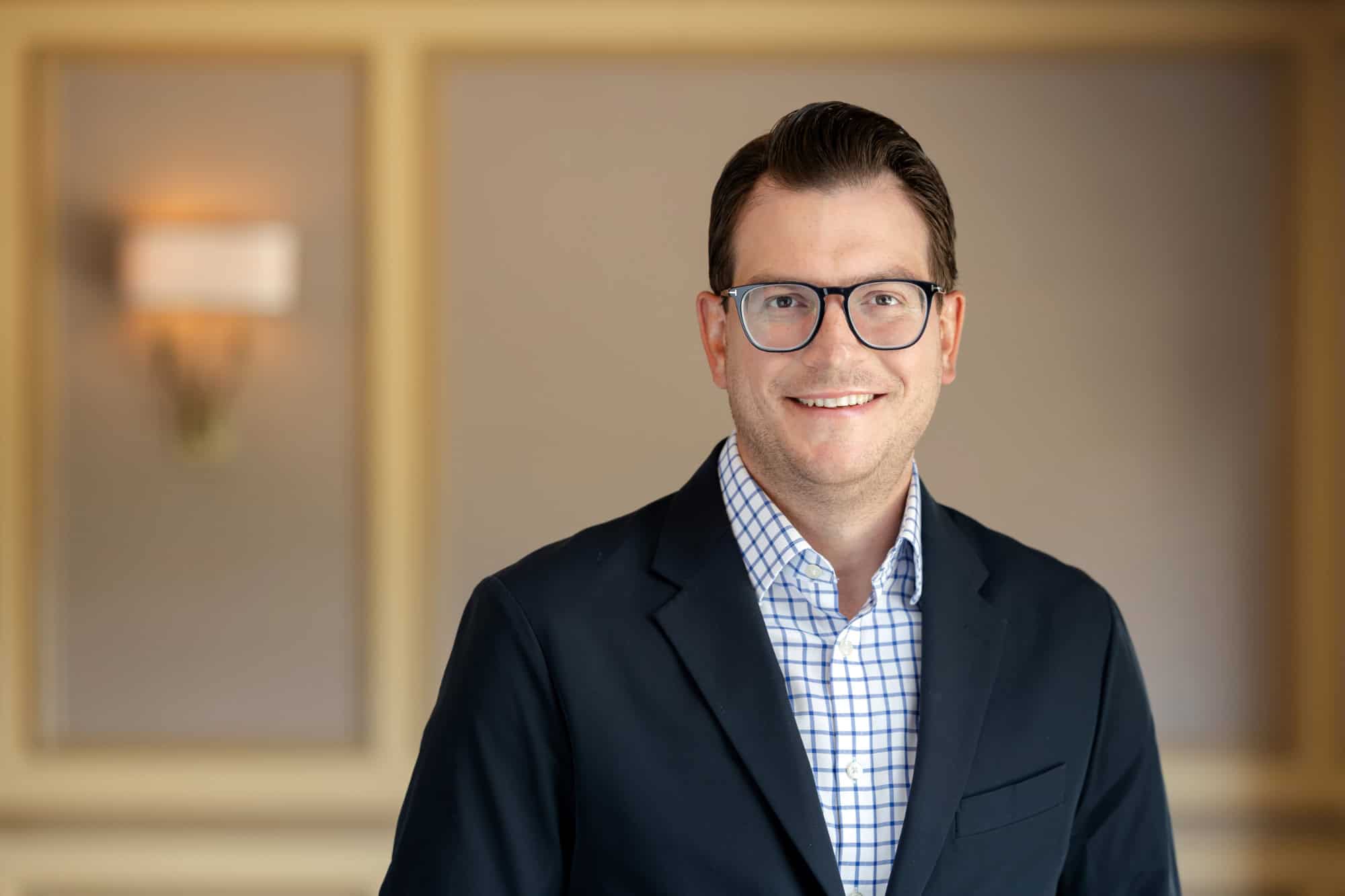 Headshot of a smiling man with dark hair and glasses, wearing a dark blazer and a blue and white checkered shirt.