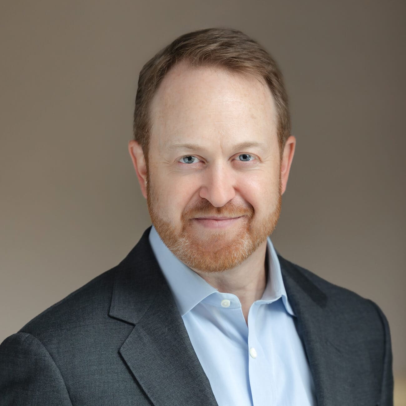 Headshot of a smiling man with light brown hair and a reddish-brown beard, wearing a dark gray suit jacket and light blue shirt.