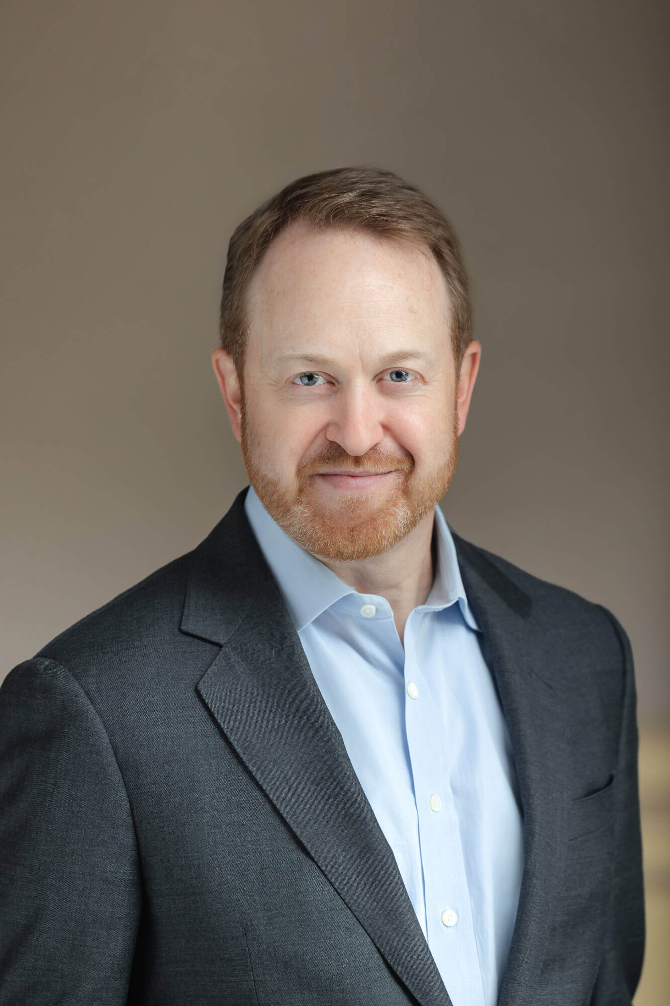 Headshot of a smiling man with light brown hair and a reddish-brown beard, wearing a dark gray suit jacket and light blue shirt.