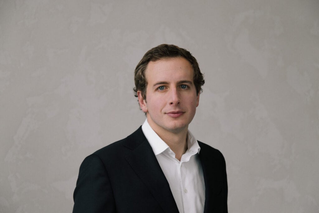 Headshot of a man with blue eyes and brown hair, wearing a black suit jacket and white shirt, against a light gray background.
