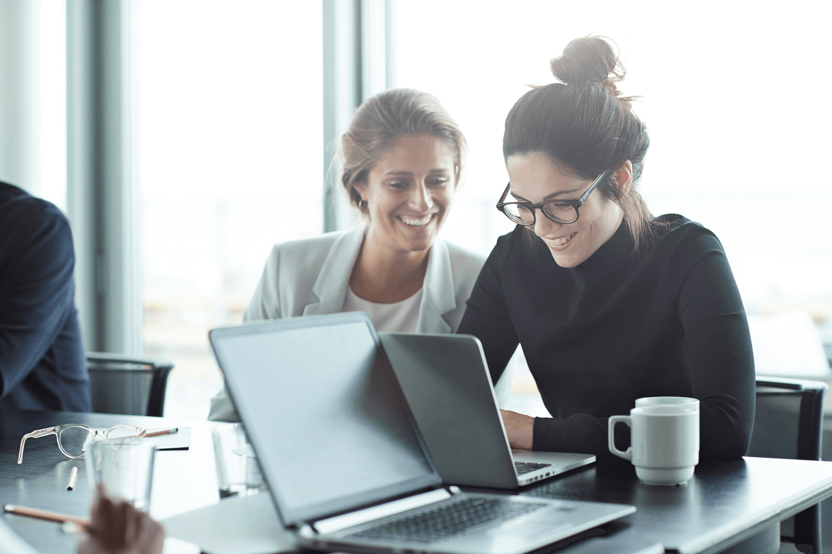Two smiling women looking at a laptop in a bright office, with a man partially visible on the left.