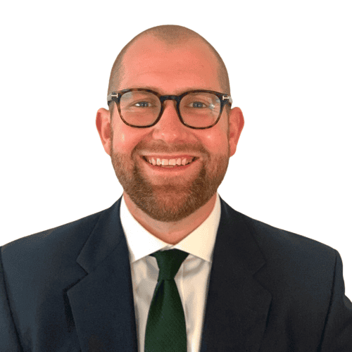 Headshot of a smiling man with a bald head, beard, and glasses, wearing a dark suit and green tie against a white background.