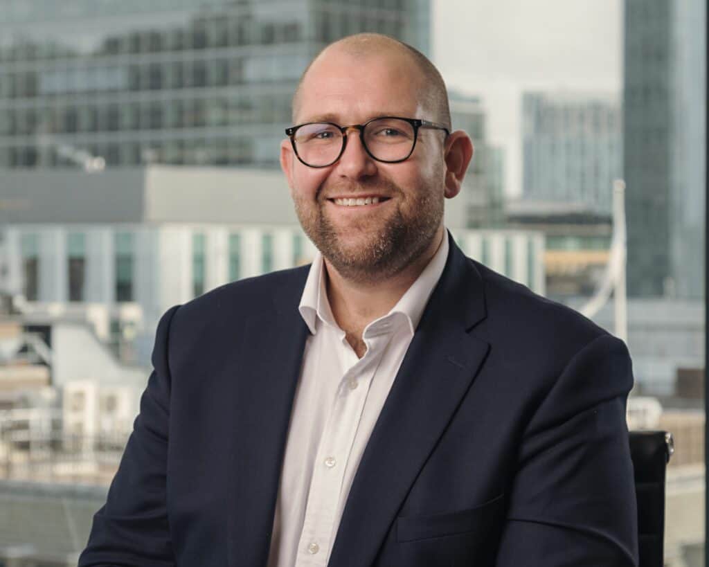 A smiling bald man with a beard and glasses, wearing a dark suit and white shirt, in front of a blurred city window.