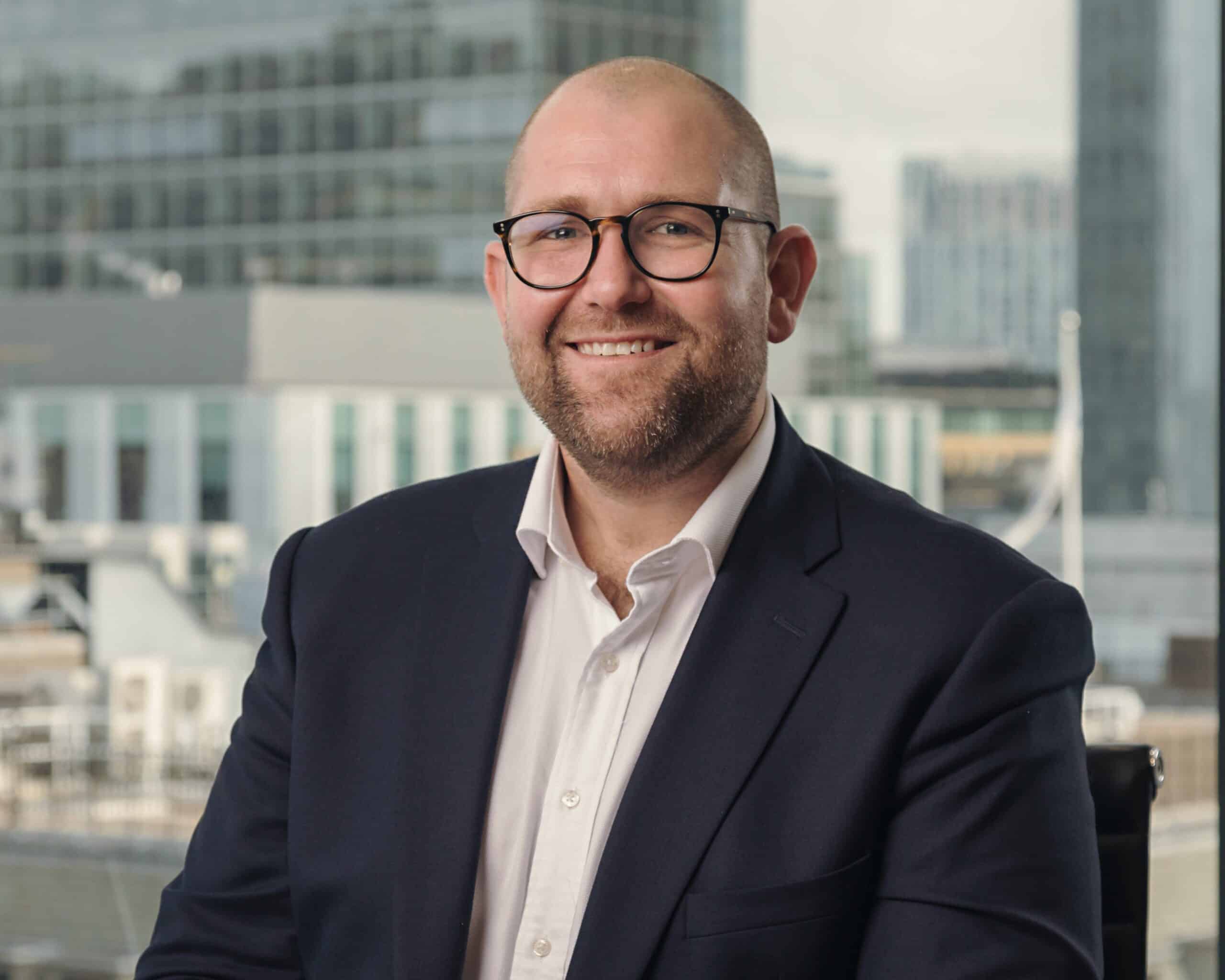 A smiling bald man with a beard and glasses, wearing a dark suit and white shirt, in front of a blurred city window.