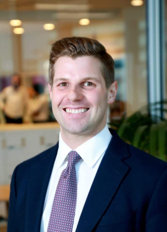 Headshot of a smiling man in a dark blue suit, white shirt, and patterned tie, with a blurred office background.