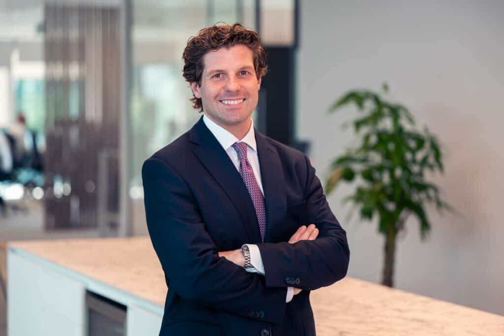 A man in a dark suit and tie, with curly hair, smiles at the camera with his arms crossed in an office.