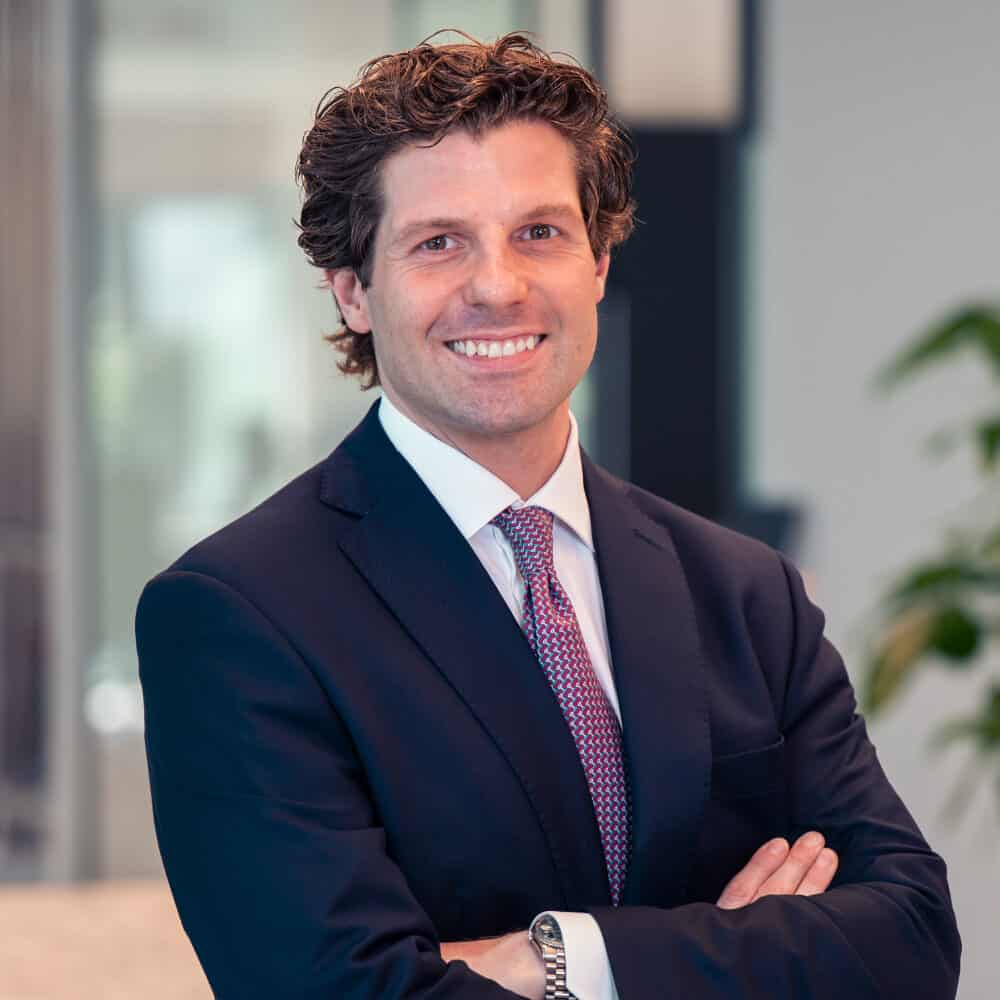 A man in a dark suit and tie, with curly hair, smiles at the camera with his arms crossed in an office.