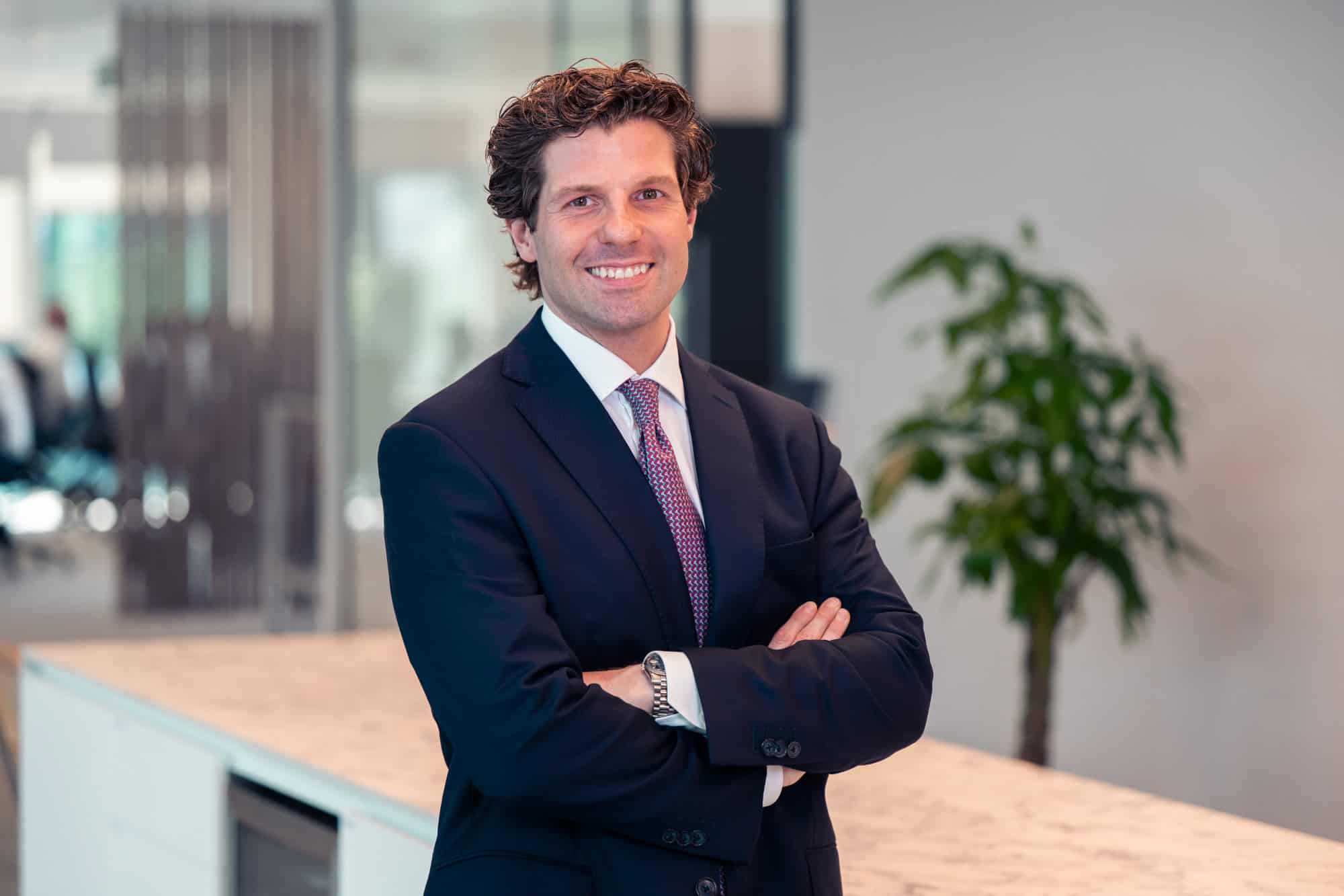 A man in a dark suit and tie, with curly hair, smiles at the camera with his arms crossed in an office.