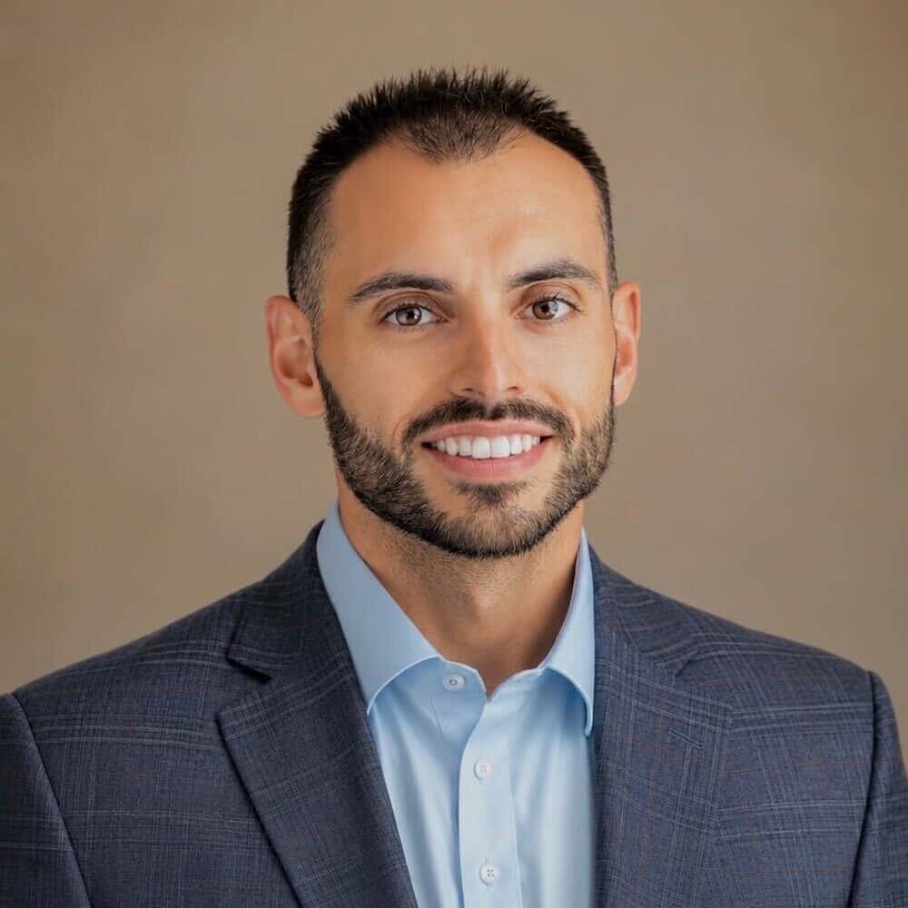 Headshot of a smiling man with dark hair and beard, wearing a light blue shirt and dark blue plaid blazer.