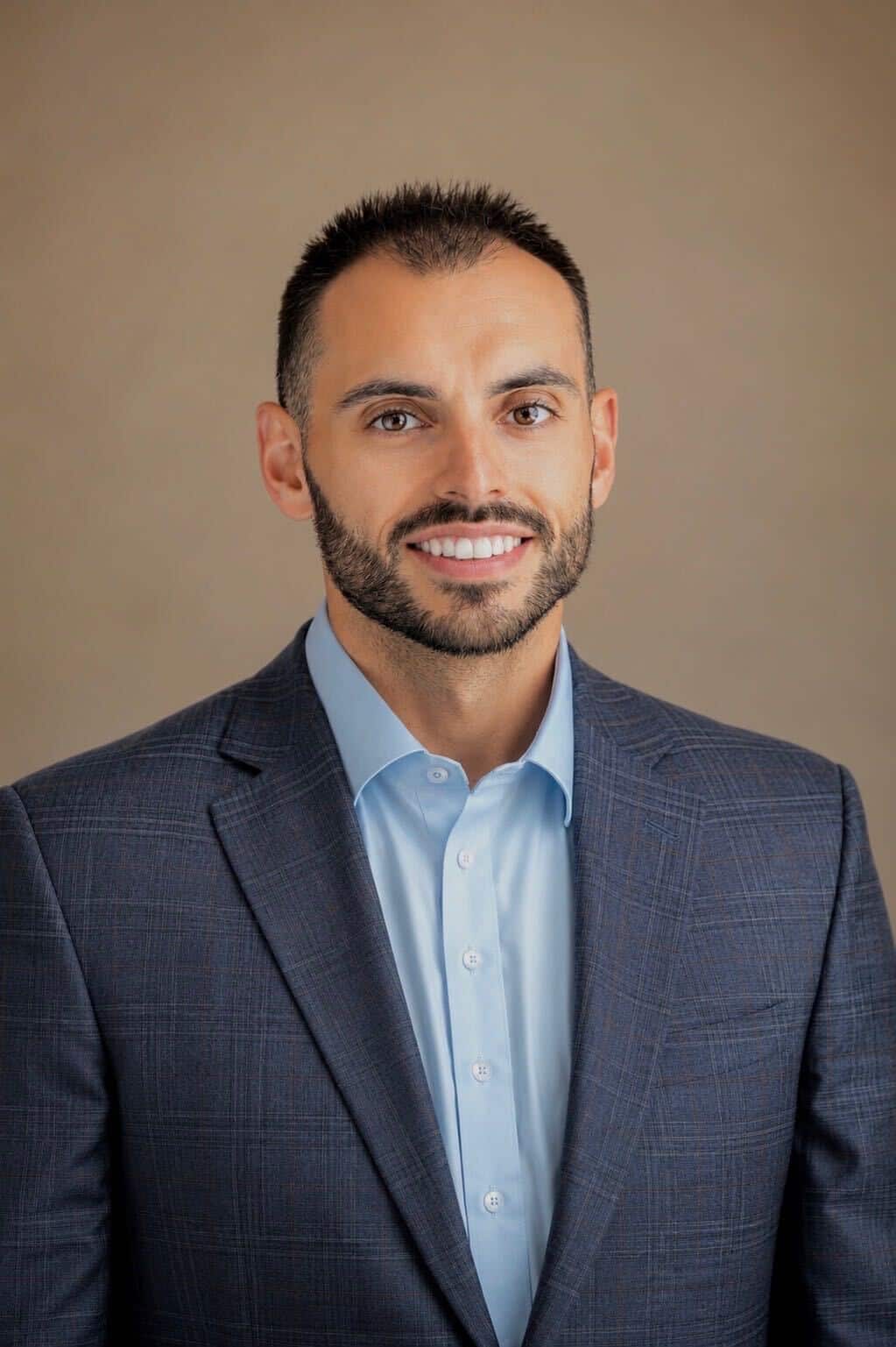 Headshot of a smiling man with dark hair and beard, wearing a light blue shirt and dark blue plaid blazer.